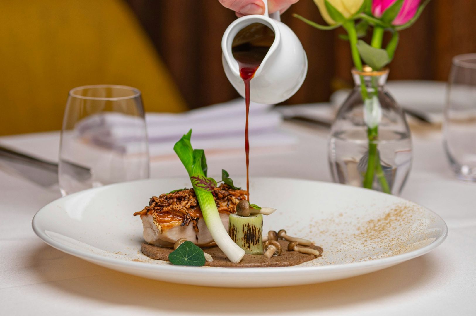 A gourmet dish on a white plate with grilled fish, greens, and sauce being elegantly poured. A stylish table setting with flowers in the background.