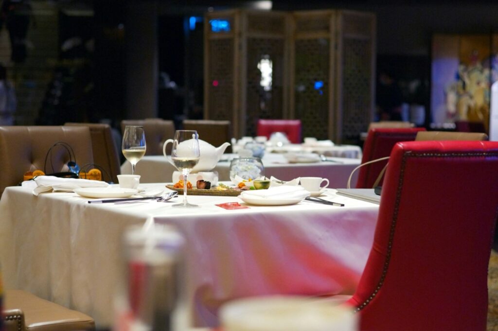 Elegant dining table set with white tablecloth, wine glasses, and decorative plates. Red and brown chairs surround the table in a dimly lit restaurant.