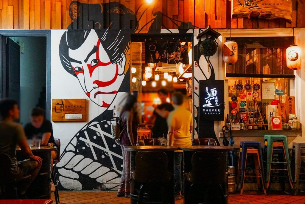 Street scene featuring a Japanese-themed restaurant with a bold mural of a kabuki actor. Warm lighting and patrons create a lively, inviting atmosphere.