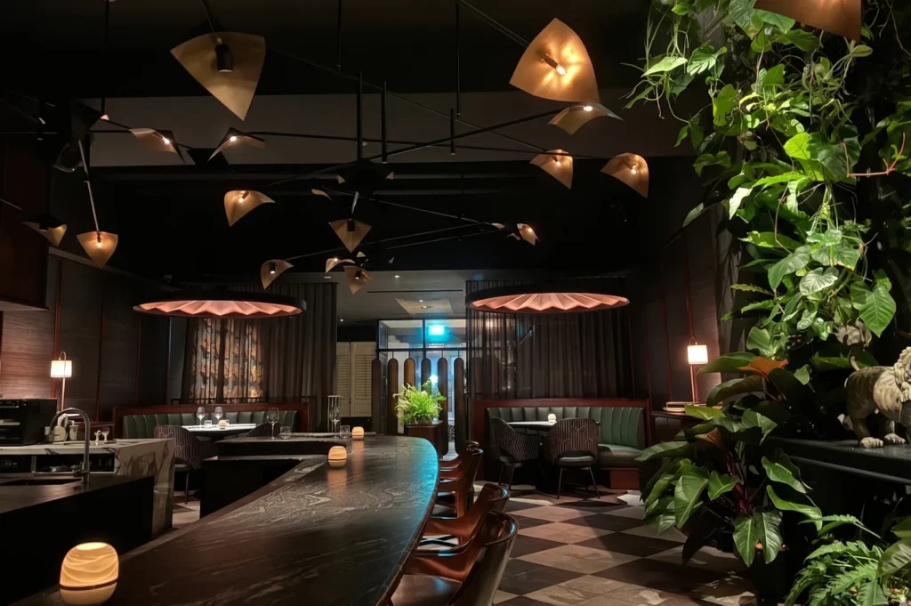 A sophisticated moody restaurant interior with dark marble counters, sculptural gold light fixtures, green leather booths, and lush indoor plants.