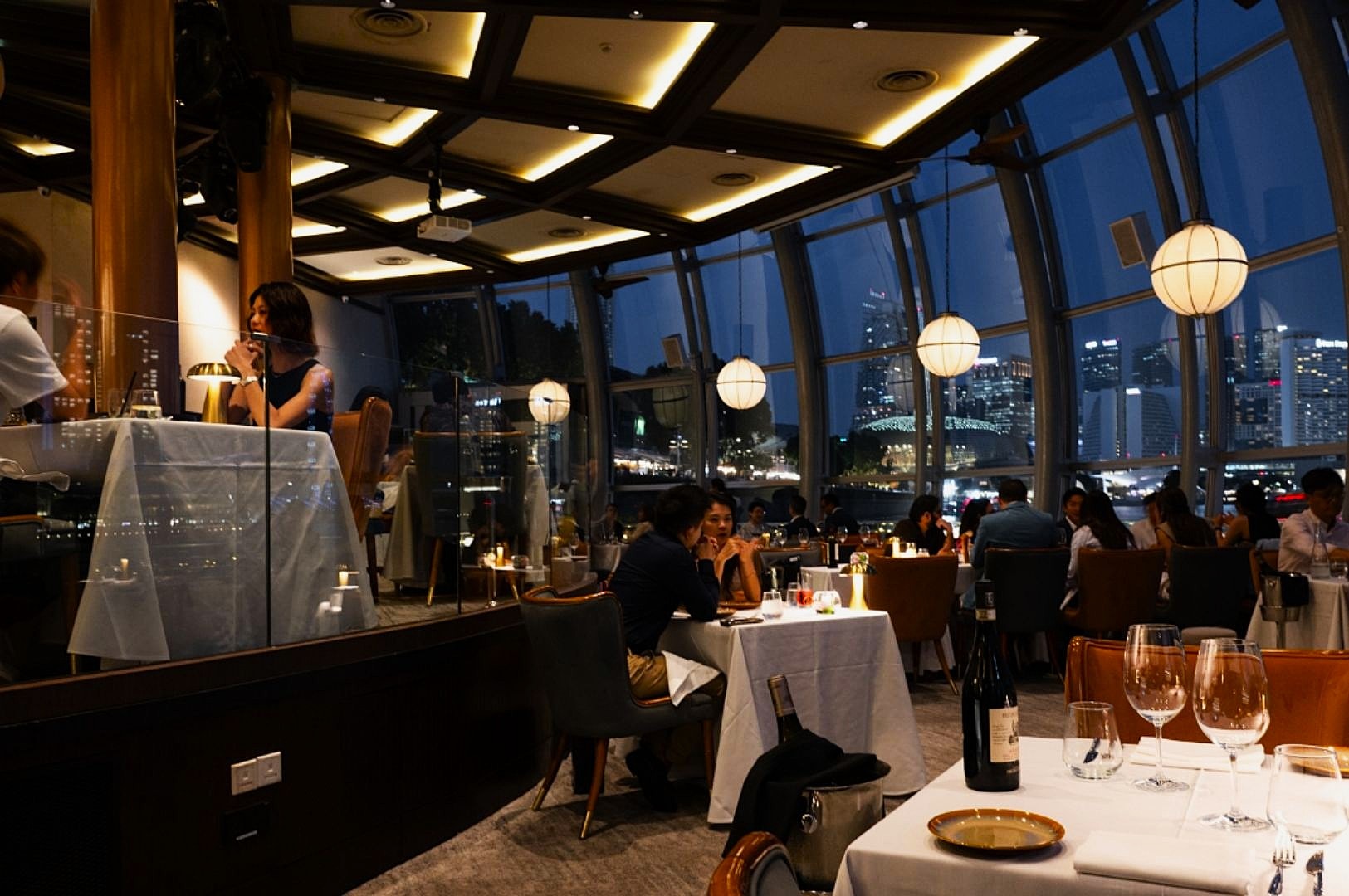 Elegant restaurant interior with large windows overlooking a cityscape at night. Guests dine at candlelit tables, creating a warm, intimate atmosphere.