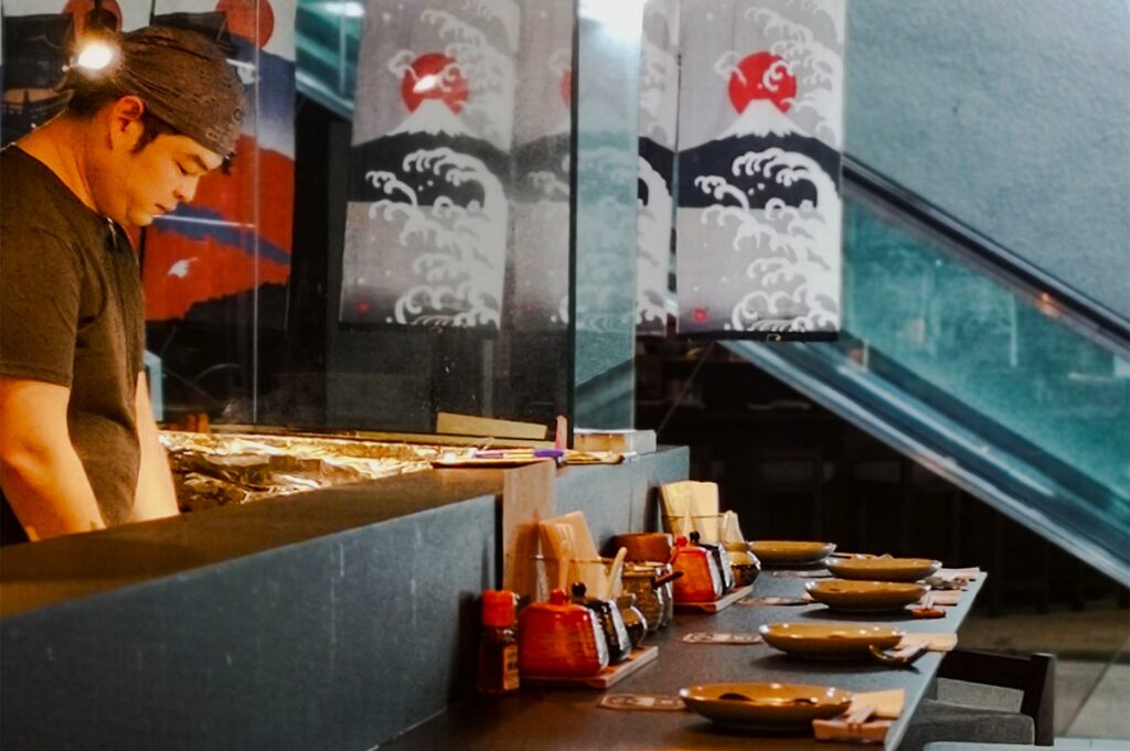 A chef in a headscarf concentrates on cooking at a sushi bar, with neatly set plates and colorful condiments. Japanese art decorates the background.