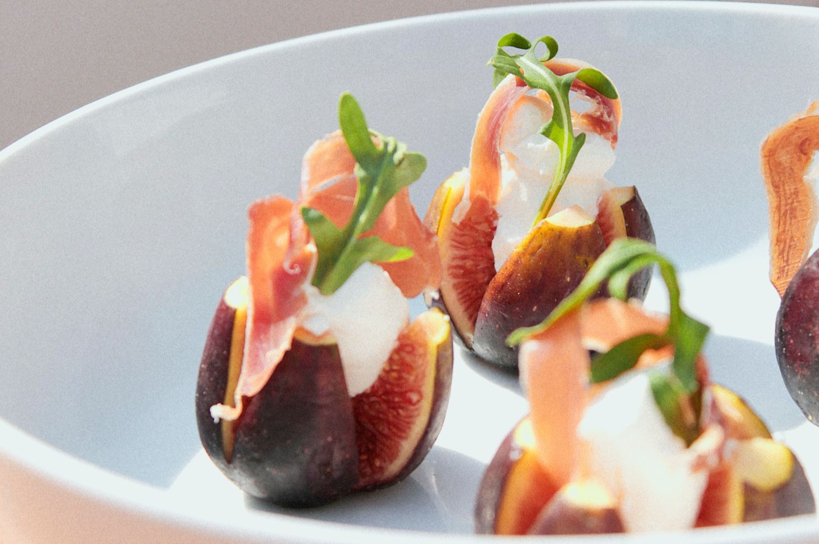 Gourmet figs in a white bowl, stuffed with creamy cheese, garnished with thin prosciutto slices and arugula. Vibrant, fresh, and elegant presentation.