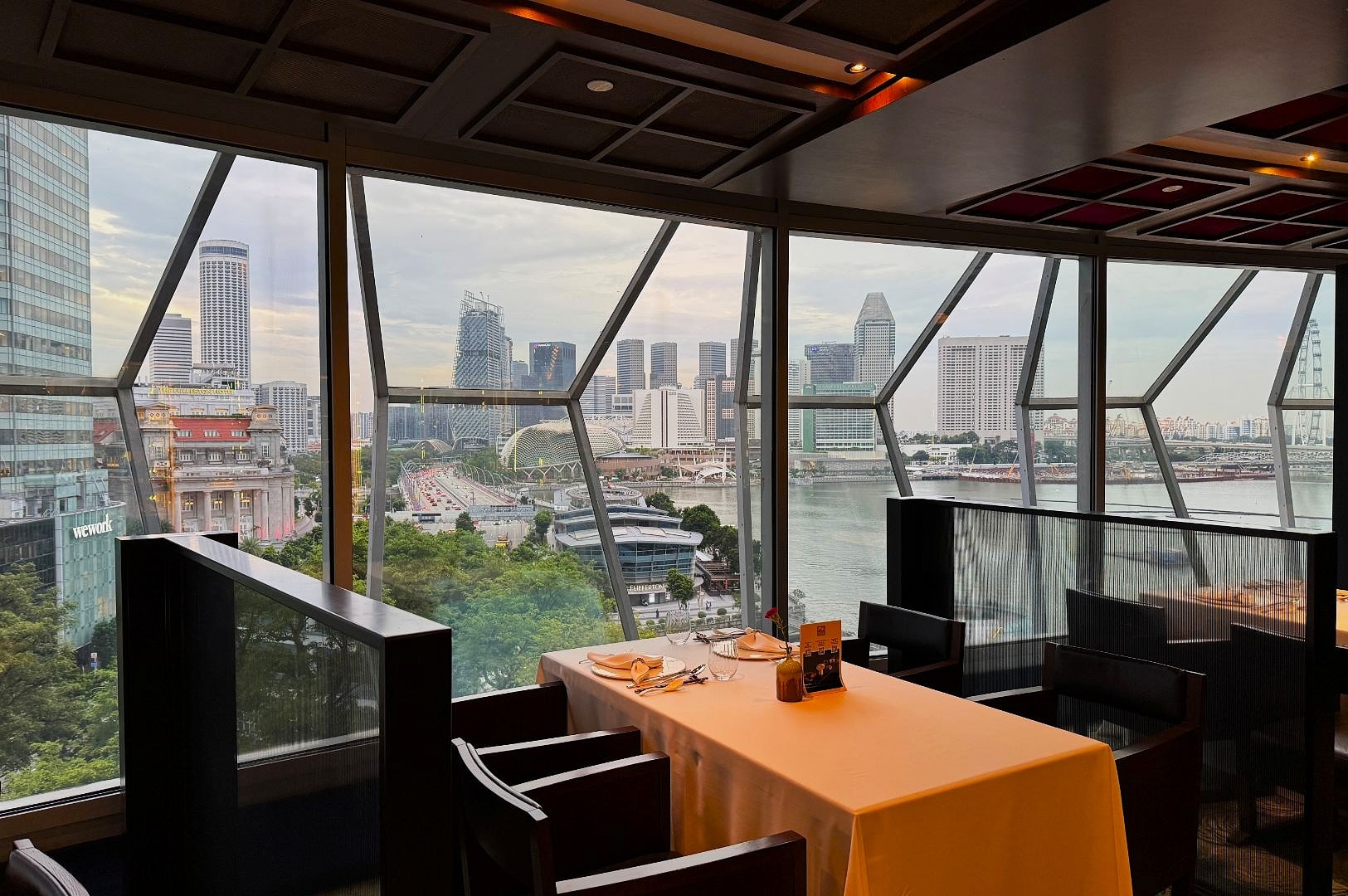 Elegant restaurant interior with a set dining table by a large window. The window offers a panoramic view of a bustling cityscape and waterfront, evoking a serene, upscale ambiance.