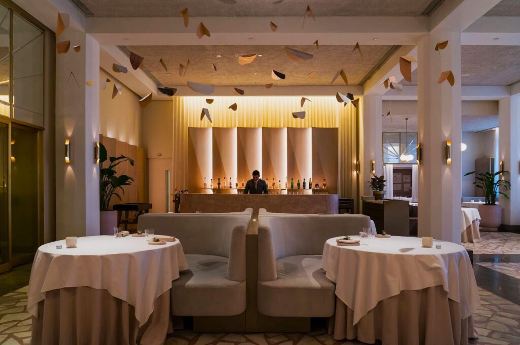 Elegant restaurant interior featuring two round tables with white tablecloths, soft lighting, and a bartender behind a stylish, backlit bar. Ceiling art adds a modern touch.