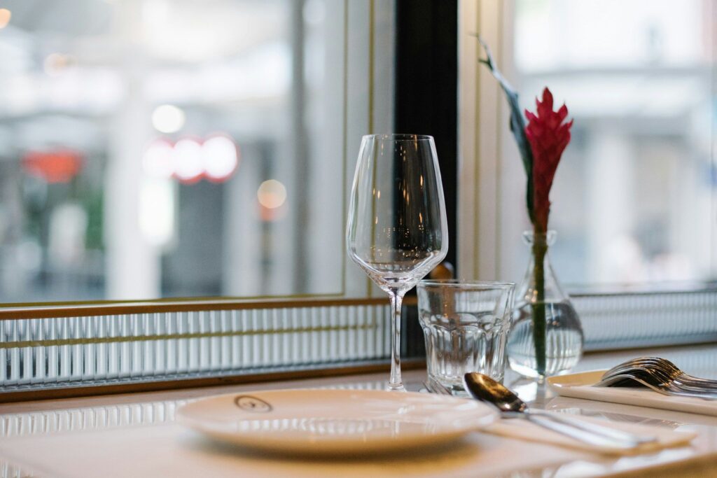A restaurant table with a wine glass, glass tumbler, and cutlery on a white cloth. A small vase with a red flower adds elegance and a cozy ambiance.
