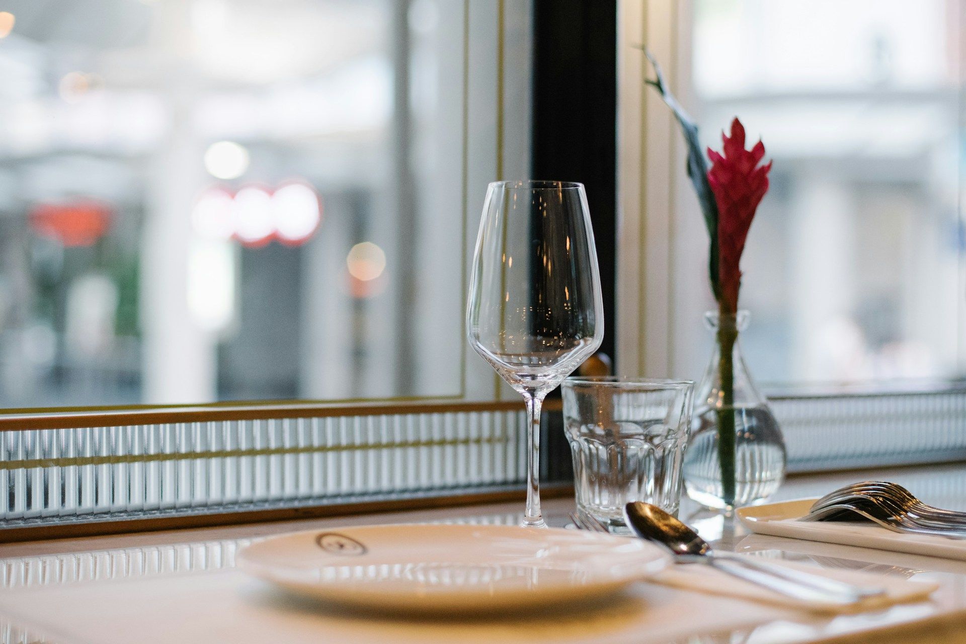 A restaurant table with a wine glass, glass tumbler, and cutlery on a white cloth. A small vase with a red flower adds elegance and a cozy ambiance.