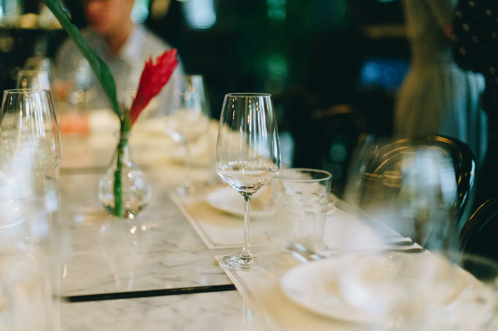 Elegant dining table set with empty wine glasses, plates, and a vase holding a red flower on a white marble surface, evoking a refined ambiance.