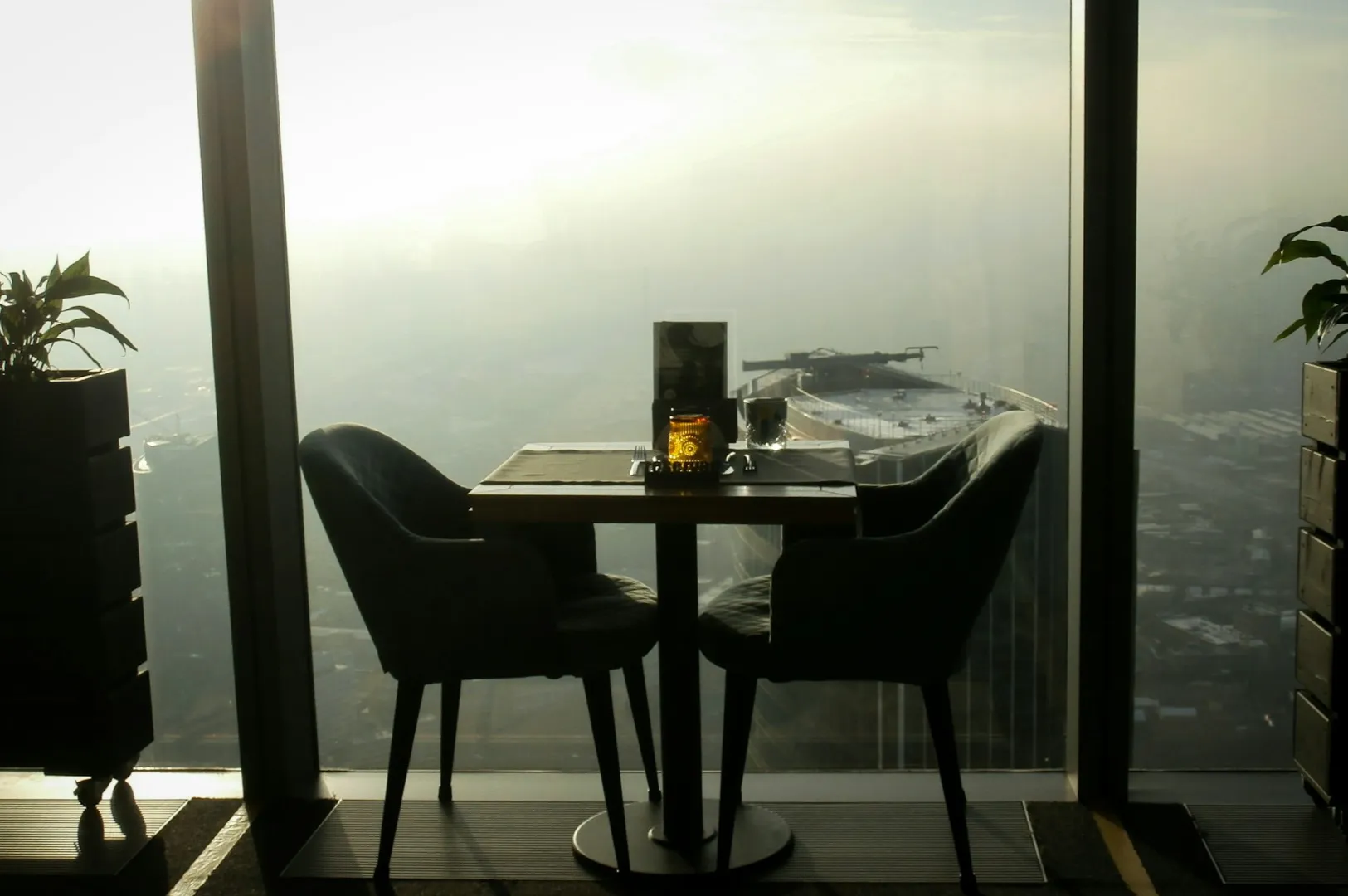 A cozy table for two by a large window overlooking a hazy urban skyline. Candlelight and plants add a warm, intimate ambiance against the bright sky.