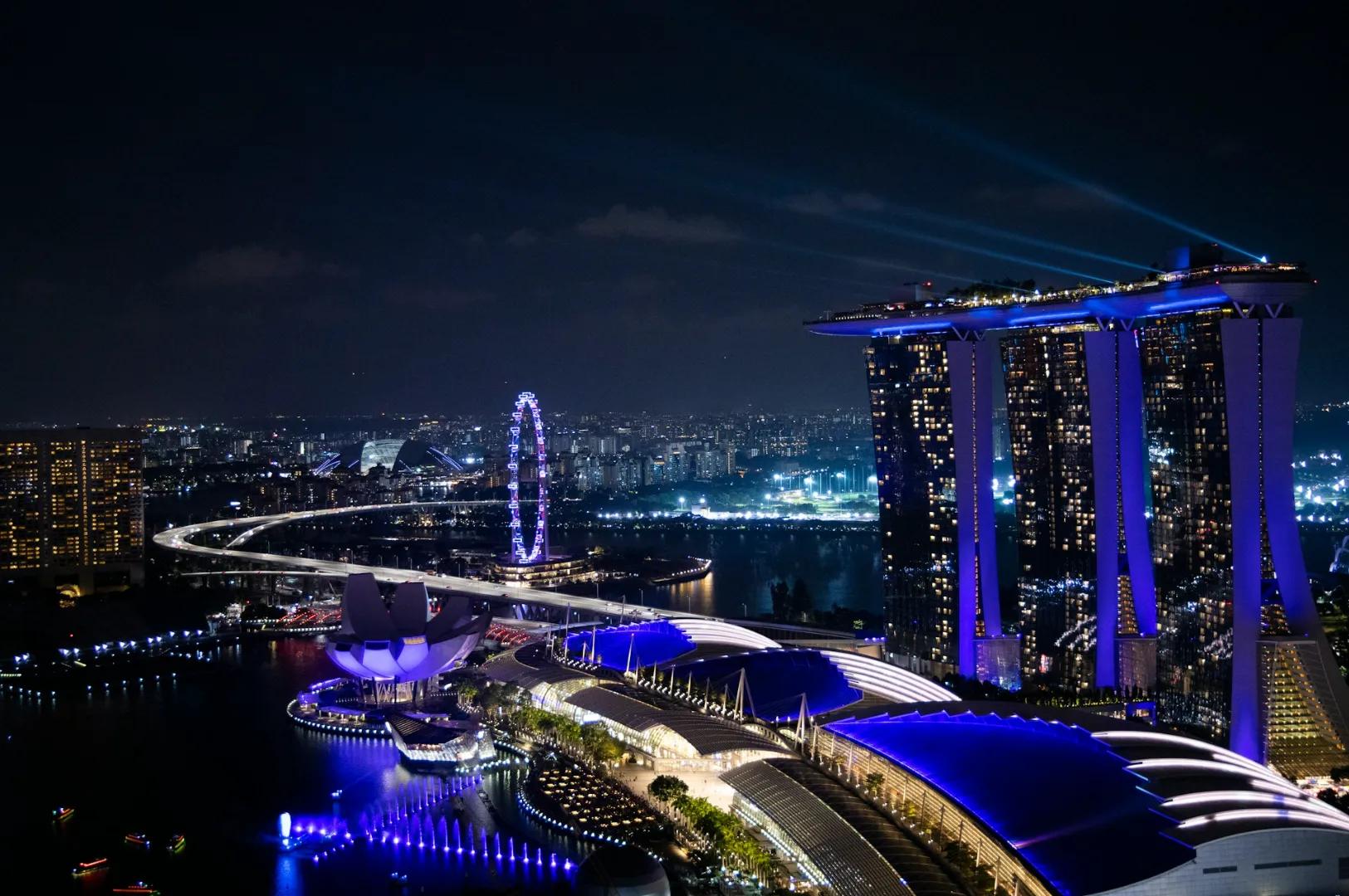 Aerial view of Singapore's Marina Bay Sands at night, with vibrant blue lights illuminating the skyline. A glowing Ferris wheel enhances the cityscape.