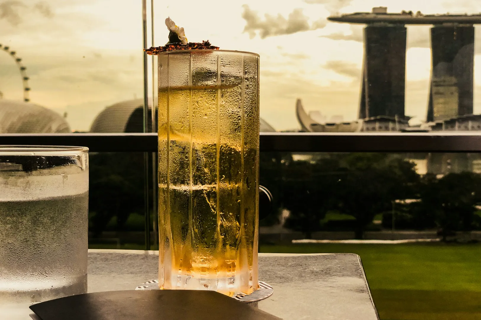 Tall, frosty cocktail on a table, overlooking Marina Bay Sands in Singapore with the iconic skyline and ferris wheel in the distance at sunset.