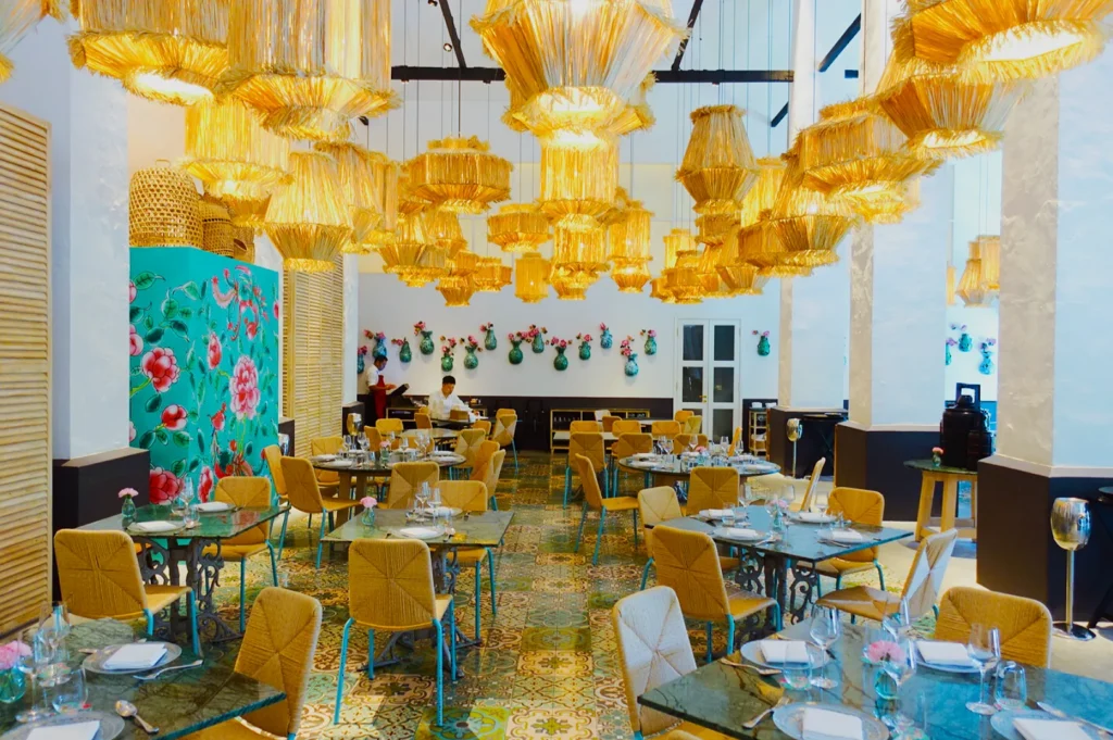 Bright, colorful restaurant with ornate yellow chandeliers, floral wall art, and vibrant tile flooring. Tables have wicker chairs, a lively ambiance.