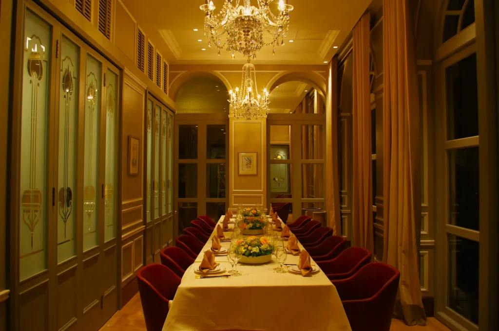 Elegant dining room with a long table set for a formal dinner, adorned with floral centerpieces. Crystal chandeliers hang above, casting a warm glow.
