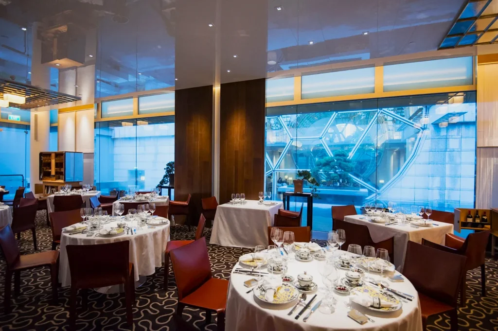 Elegant restaurant with round tables set for fine dining, featuring white tablecloths, wine glasses, and patterned china. Large window offers a blue-lit city view, creating a luxurious and serene ambiance.