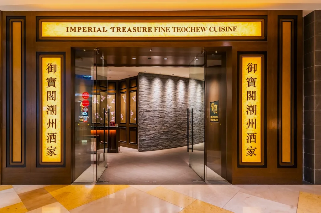 Entrance of "Imperial Treasure Fine Teochew Cuisine" restaurant with glass doors, warm lighting, wood and stone decor, creating an elegant, inviting atmosphere.