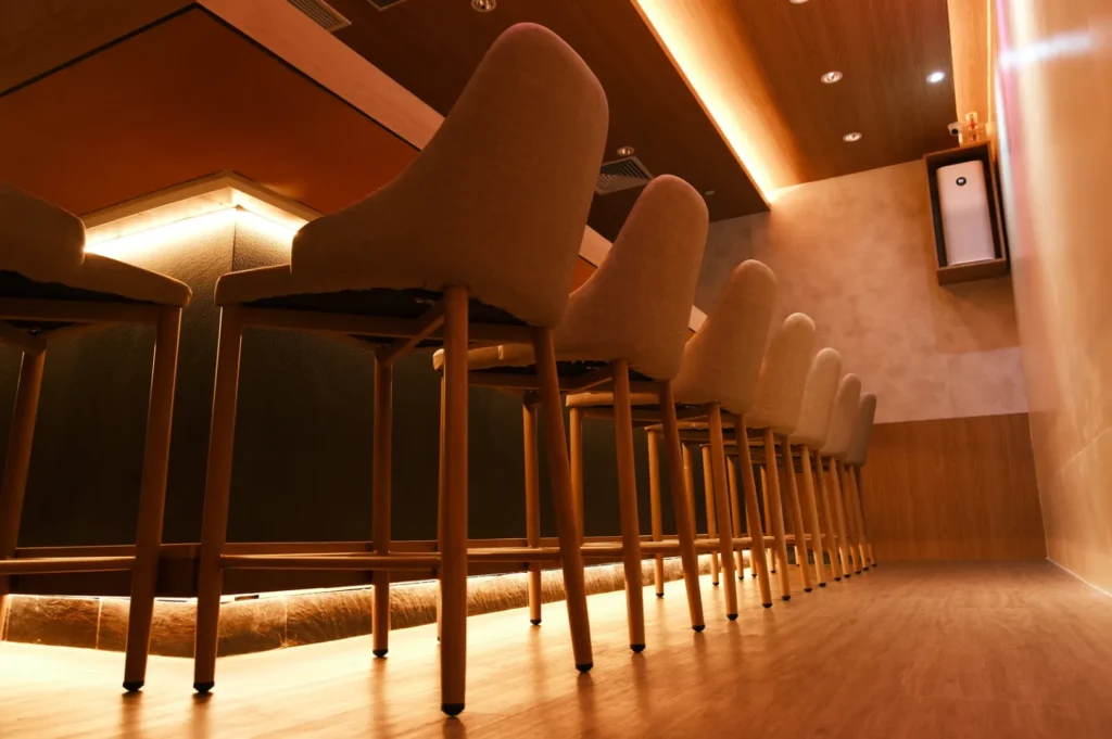Modern bar interior with a row of cushioned wooden chairs under a sleek, dimly-lit counter. Warm lighting and wooden walls create a cozy atmosphere.