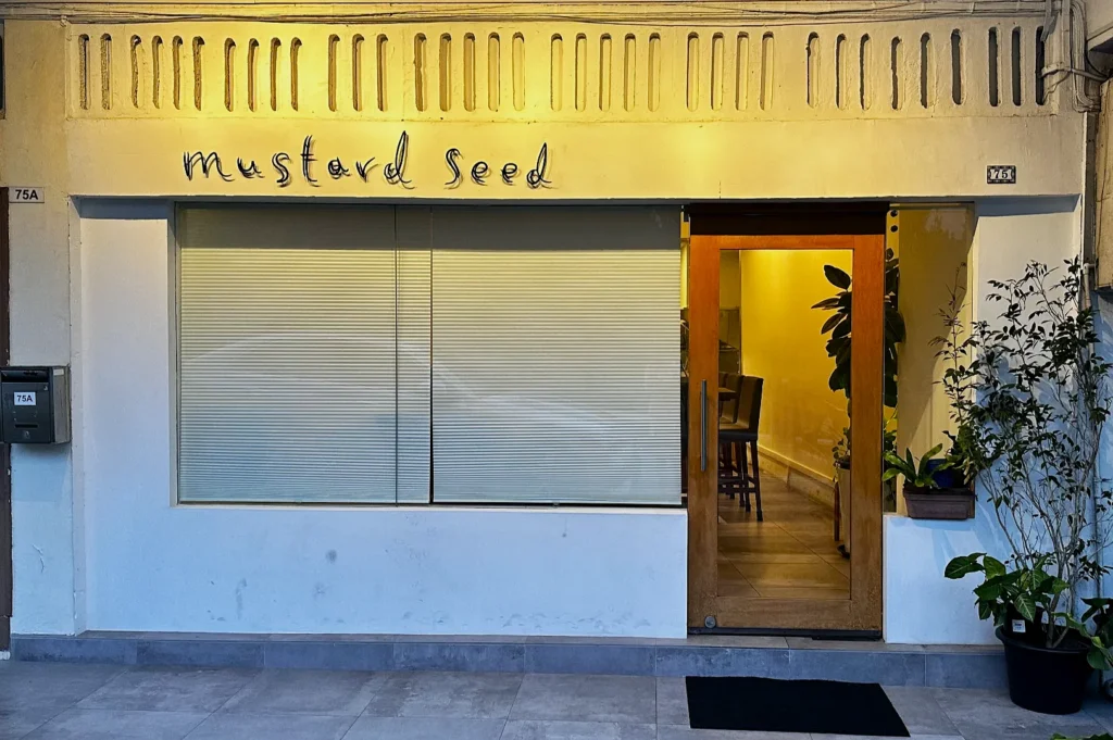 Storefront labeled "mustard seed" with closed blinds and a wooden door. Modern, minimalist ambiance with plants flanking the entrance.