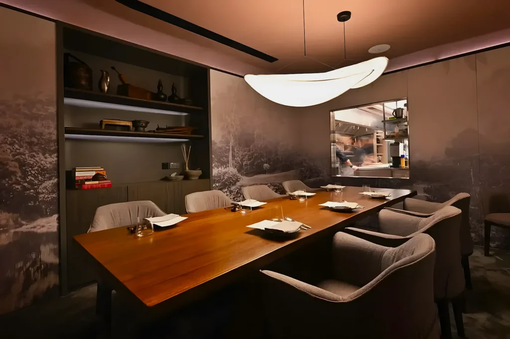 Elegant dining room with a long wooden table, plush gray chairs, and soft ambient lighting. Aesthetic wall art and open kitchen window create a cozy, refined atmosphere.