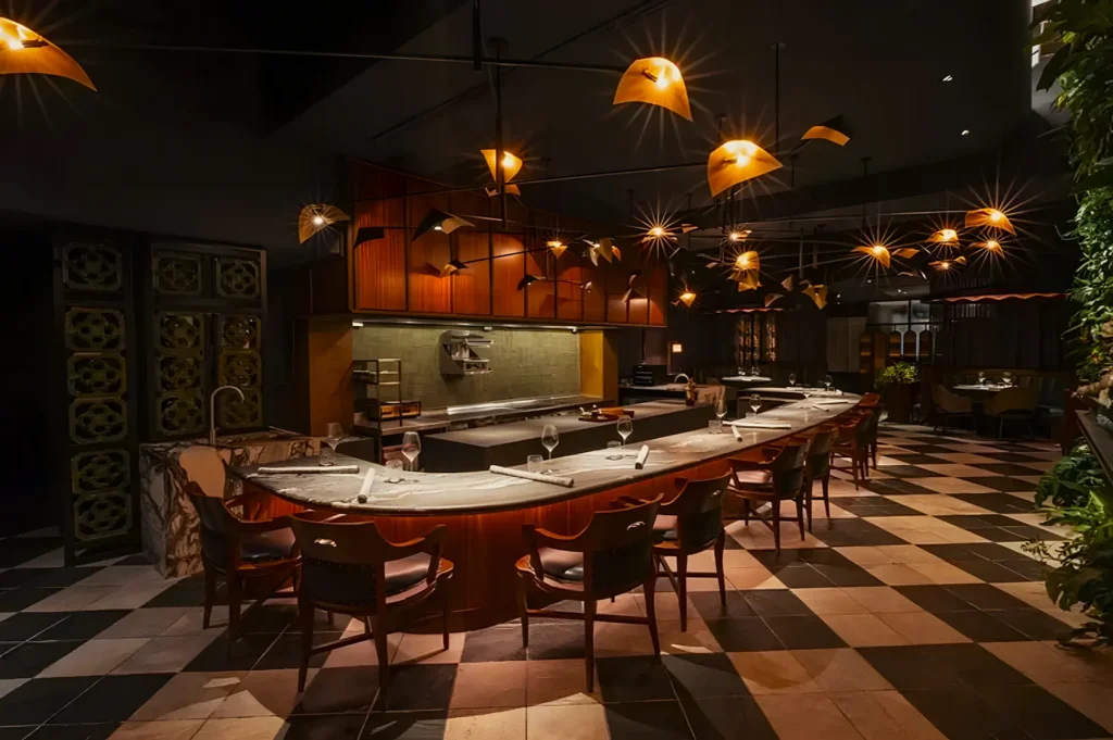 Stylish restaurant interior with a curved bar, elegant wooden chairs, and warm lighting. Checkered floor and lush greenery enhance the cozy ambiance.