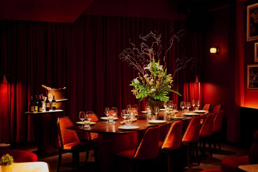 A candlelit private dining Singapore room with a long wooden table set for twelve, elegant glassware, and a tall lush floral centerpiece beneath warm red lighting.