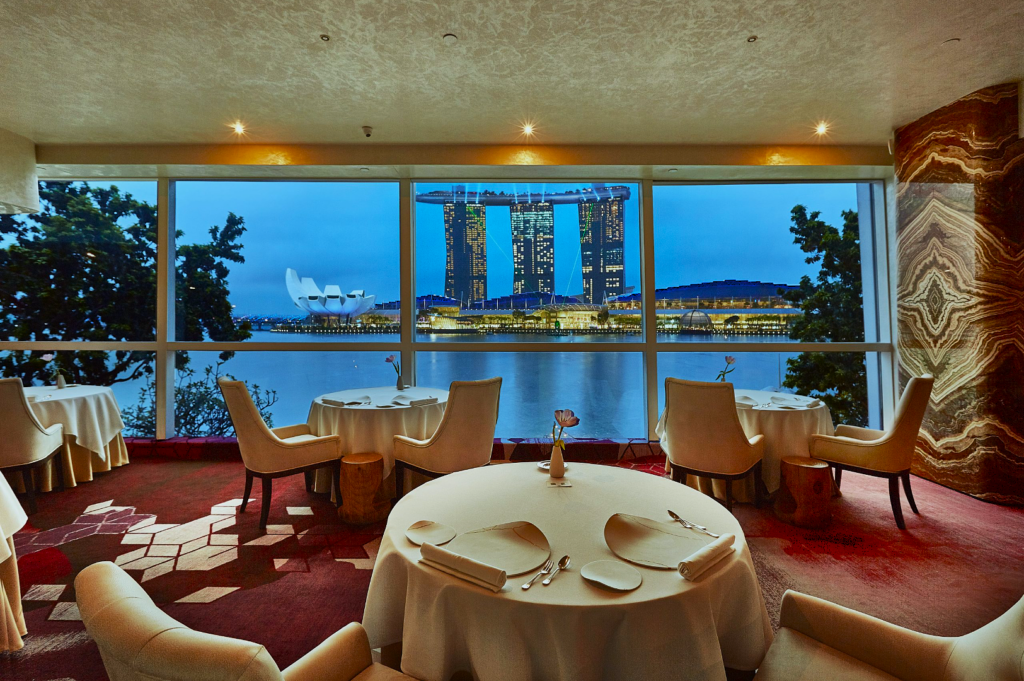 Elegant restaurant interior overlooking Marina Bay Sands through large windows, set with round tables, white tablecloths, and plush chairs at sunset.