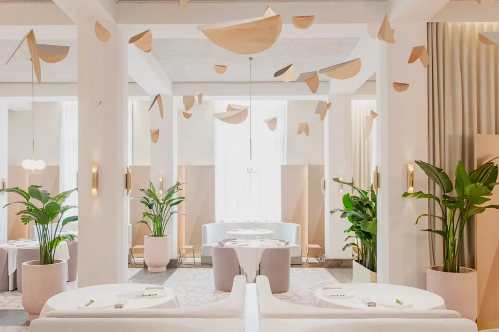 Elegant, modern restaurant interior with white and pastel tones. Suspended wooden shapes, lush green plants, and soft lighting create a serene ambiance.