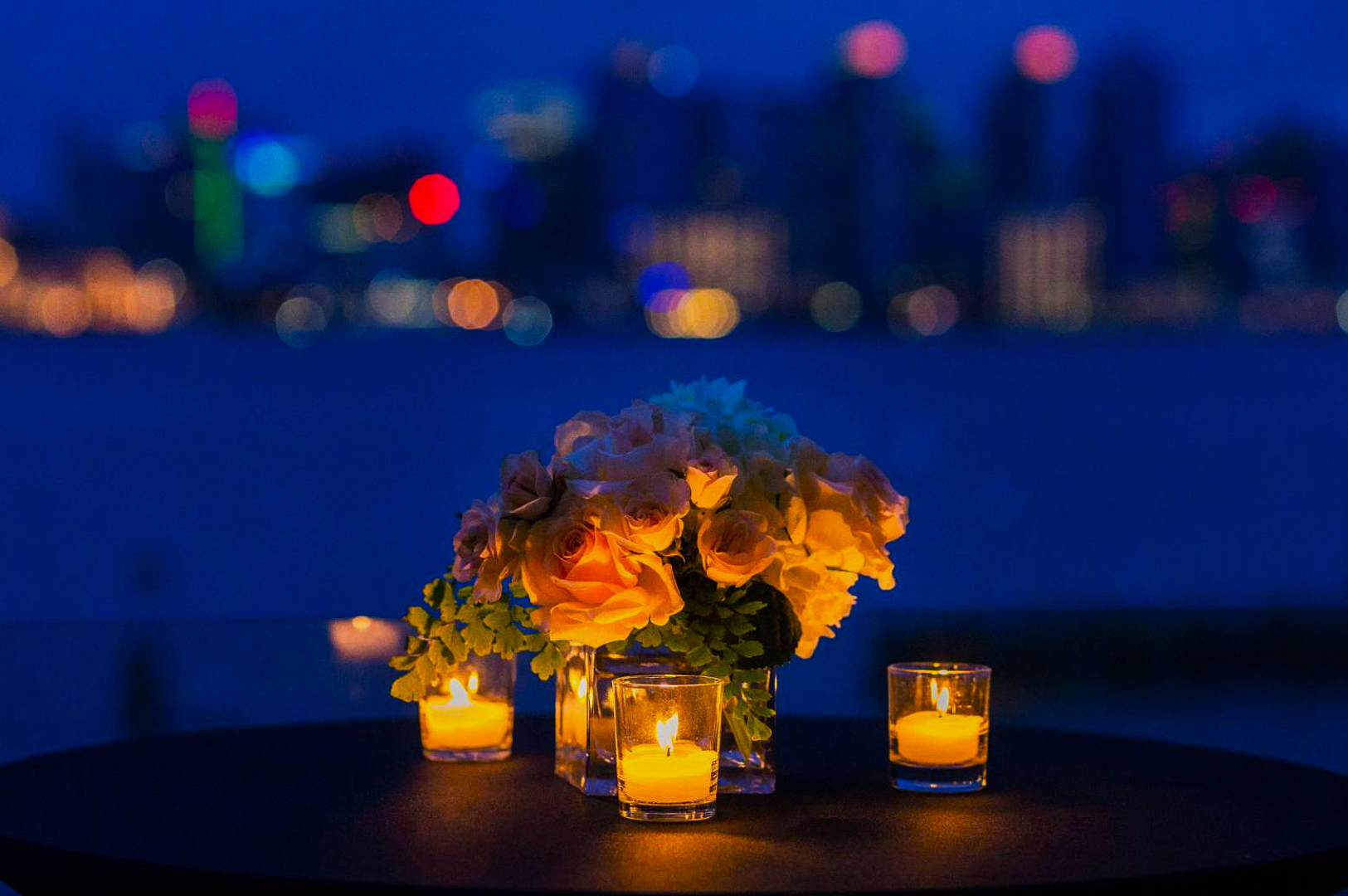 A bouquet of pale roses with greenery and three lit candles on a table. Blurred cityscape lights create a romantic evening ambiance in the background.