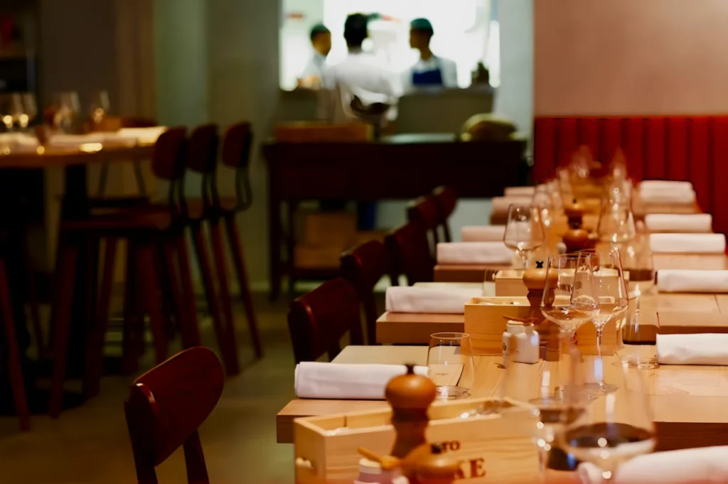 Elegant restaurant setting with wooden tables, neatly arranged glasses, and white napkins. Warm lighting creates a welcoming, cozy ambiance. Chefs are blurred in the background, adding a sense of activity.