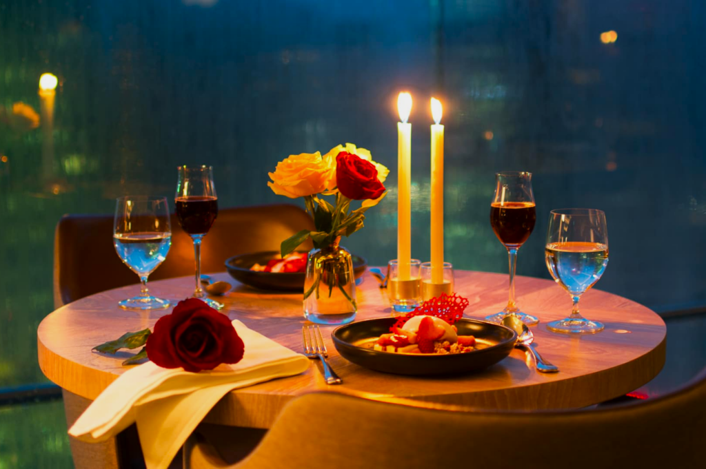 A romantic candlelit dinner setup with two plates of dessert, wine glasses, and red roses on a round table. Warm light creates an intimate ambiance.