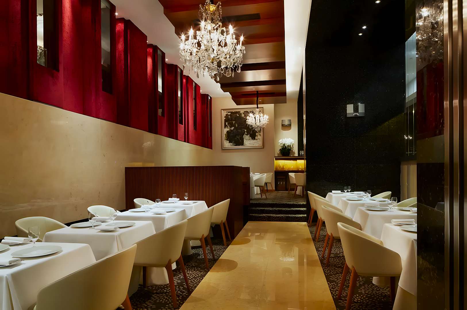Elegant restaurant with white tablecloths, plush cream chairs, and chandeliers. Red panels on walls create a warm ambiance. A painting and plant adorn the back wall.