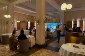 Elegant restaurant interior with round tables, white tablecloths, and soft lighting. People dine and chat, while waiters serve with attentiveness.