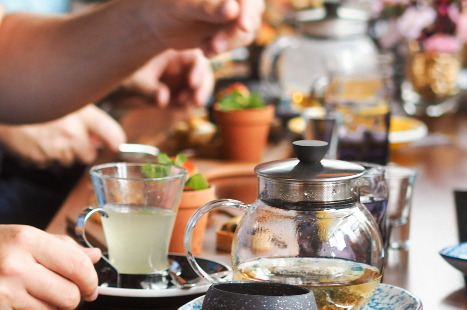A cozy tea gathering with a glass teapot and herbal tea-filled cup on a wooden table. Blurred hands reach for snacks, creating a warm, inviting atmosphere.