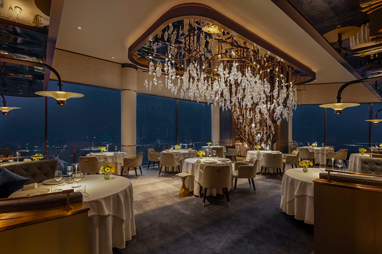 Elegant restaurant interior with round tables draped in white, overlooking a cityscape at night. A crystal chandelier adds luxury and sophistication.