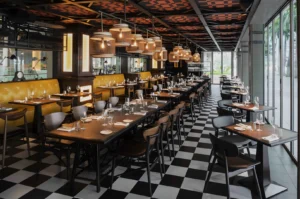 A modern restaurant interior with a warm ambiance, featuring patterned ceiling, pendant lights, checkerboard floor, wooden tables, and yellow cushioned booths.