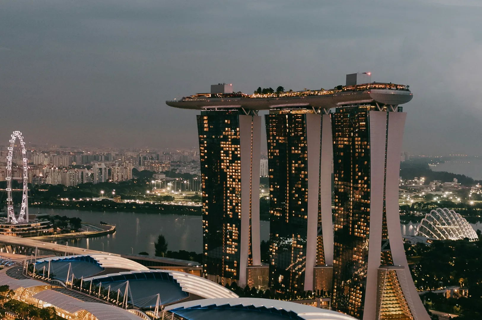 Aerial view of Marina Bay Sands in Singapore at dusk, with illuminated windows and rooftop gardens. The Singapore Flyer is visible on the left.