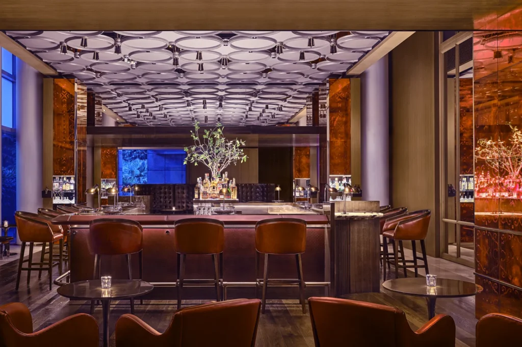 Wide-angle interior shot of a luxury hotel cocktail bar featuring a central marble bar counter, burgundy leather bar stools, mirrored wall panels, and sculptural ceiling lighting, photographed