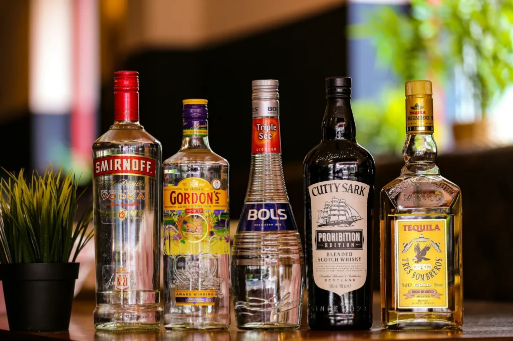 Five liquor bottles, including vodka, gin, and whisky, arranged on a wooden table next to a small potted plant, with a blurred background.