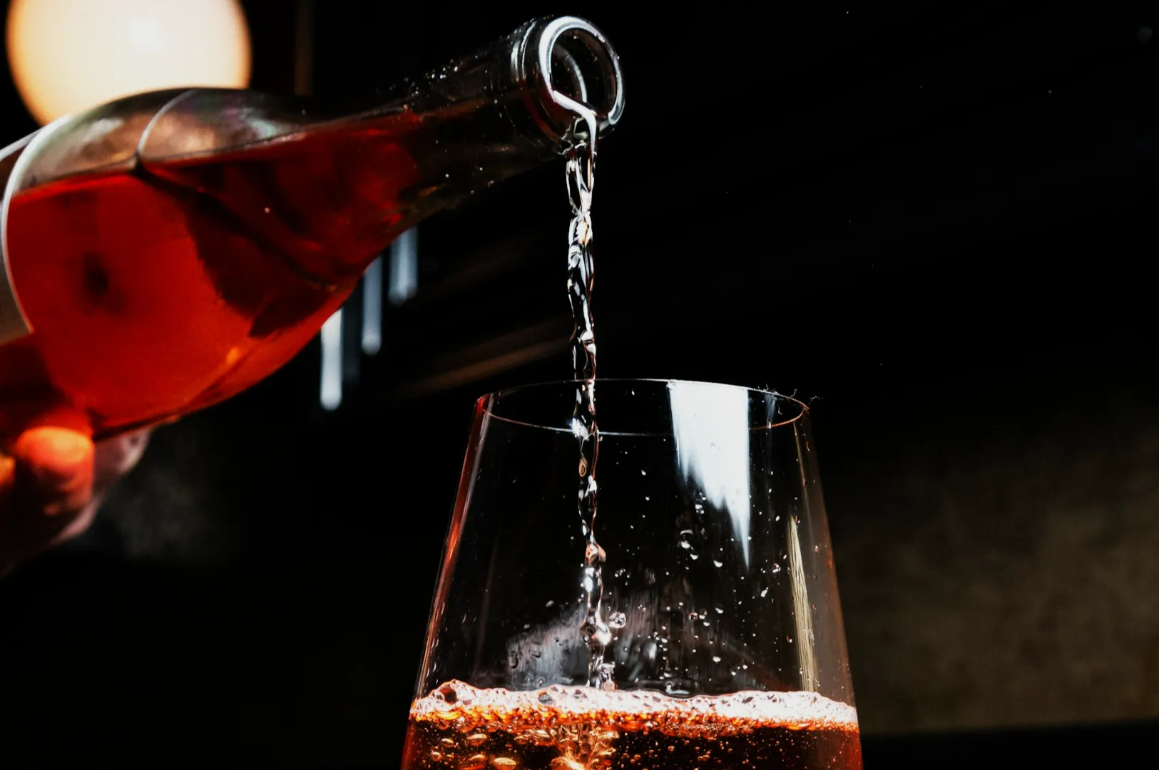 A stream of rosé wine pours from a bottle into a clear glass, with bubbles visible. The background is softly lit, creating an elegant and inviting mood.