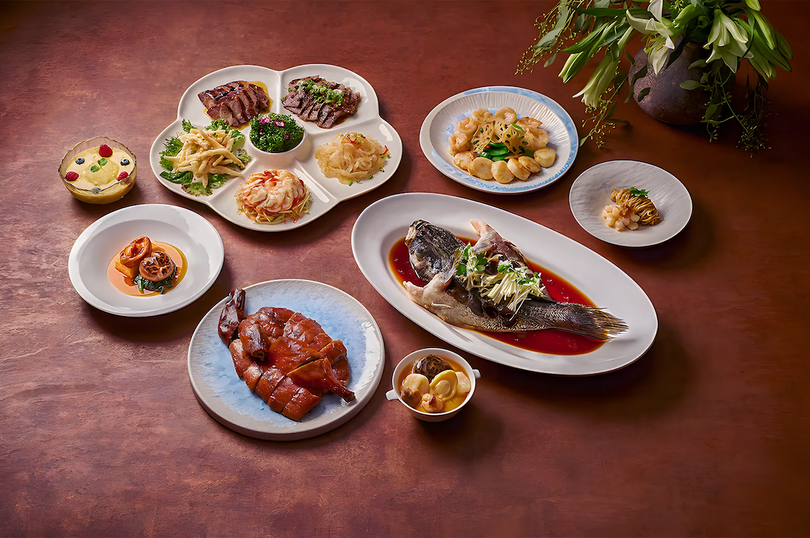 A sumptuous spread of Asian cuisine on a brown table. Includes steamed whole fish in sauce, sliced duck, assorted appetizers, dessert, and a green plant centerpiece.