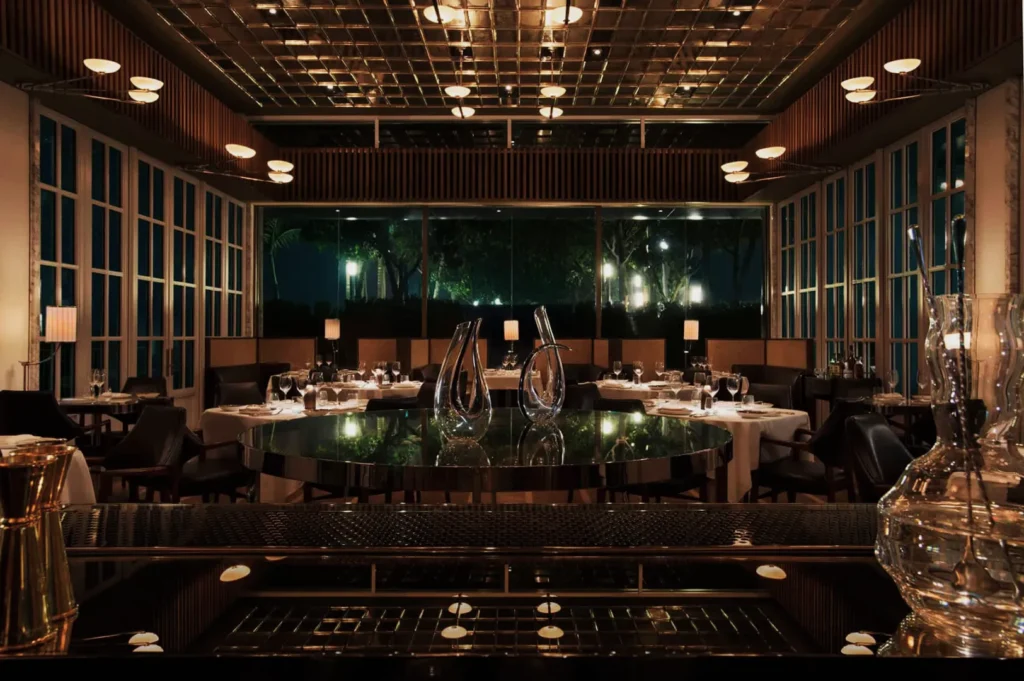 Elegant restaurant interior at night, featuring a central reflective table, ambient lighting, large windows with tree views, and neatly set tables.
