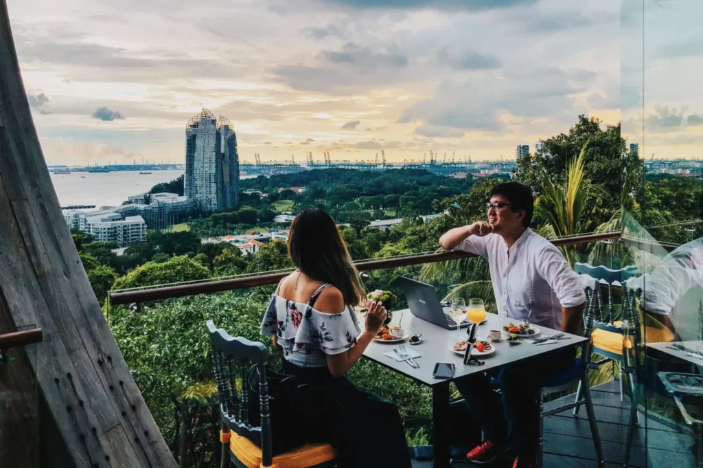 A couple enjoys an alfresco dinner at a balcony table overlooking a lush landscape and distant city skyline at sunset, creating a serene atmosphere.
