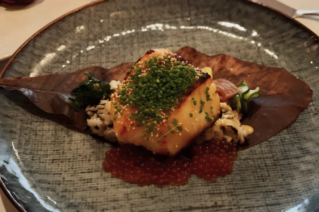 A gourmet dish featuring a seared fish fillet topped with finely chopped green herbs on a brown leaf, surrounded by rice, seaweed, and orange roe.