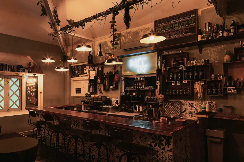 Cozy bar with wooden counter, stools, and warm hanging lights. Shelves filled with bottles line the wall, and a neon sign reads "Don't Try." Lush plants add a relaxed atmosphere.