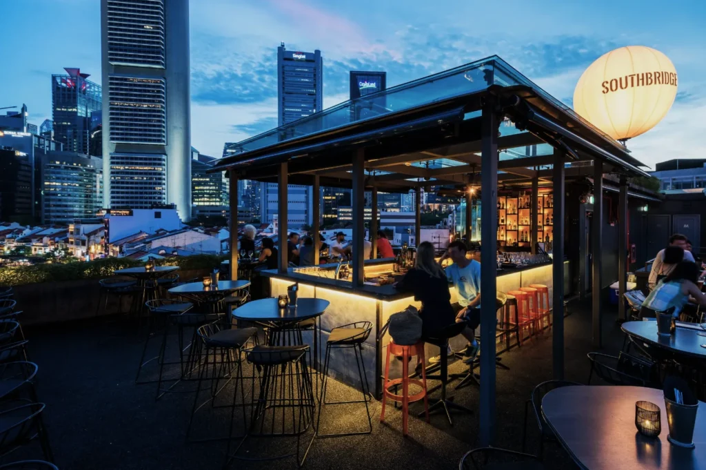 Rooftop bar at twilight with a city skyline backdrop, featuring round tables, glowing lanterns, and people enjoying drinks. Atmosphere is relaxed and vibrant.