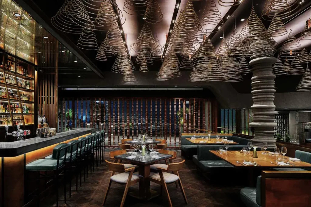 Chic restaurant interior with spiral light fixtures, modern bar, and wooden tables set for dining. Warm lighting creates an inviting, sophisticated ambiance.