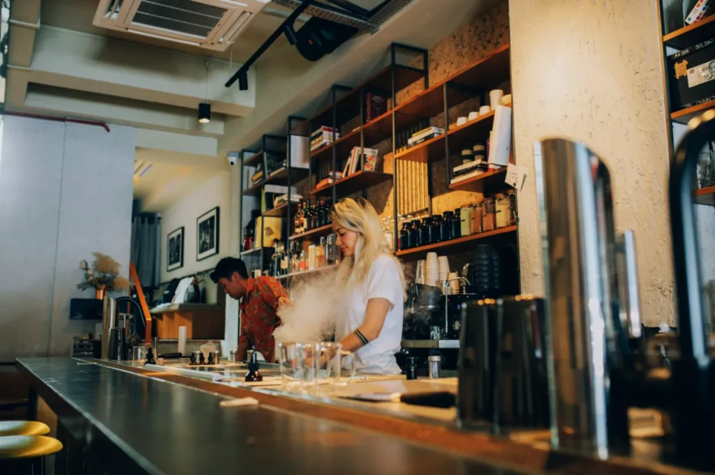 Baristas in a modern café prepare drinks behind a counter, surrounded by shelves of cups and bottles. A cozy, bustling atmosphere is conveyed.