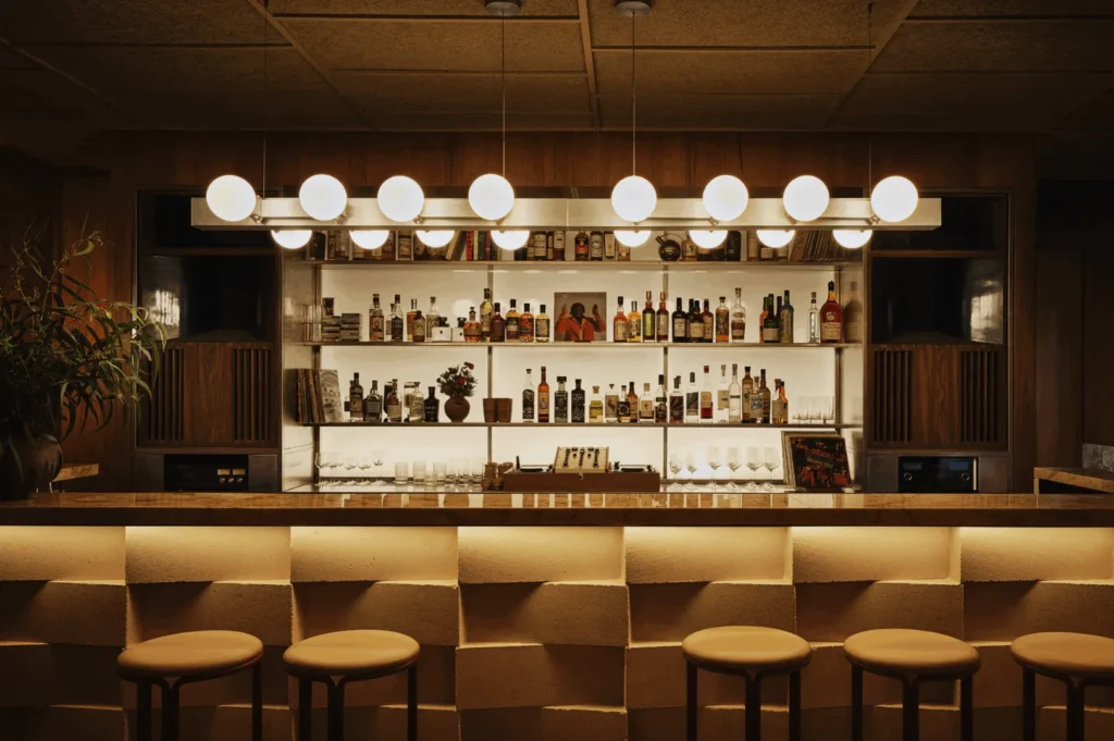A cozy, dimly lit bar with a modern design. Round lights hang above a textured counter with stools. Shelves display bottles and decor, creating a warm ambiance.