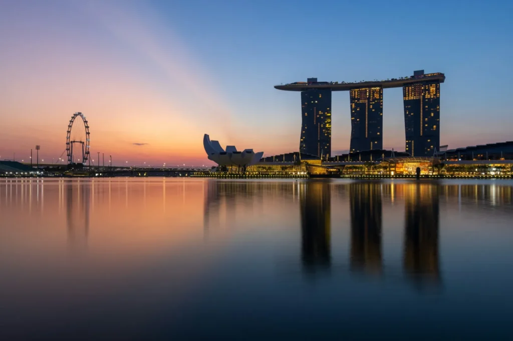 Sunset over Singapore's Marina Bay with skyline reflections on water. Marina Bay Sands and Singapore Flyer silhouette against vibrant orange and blue sky.