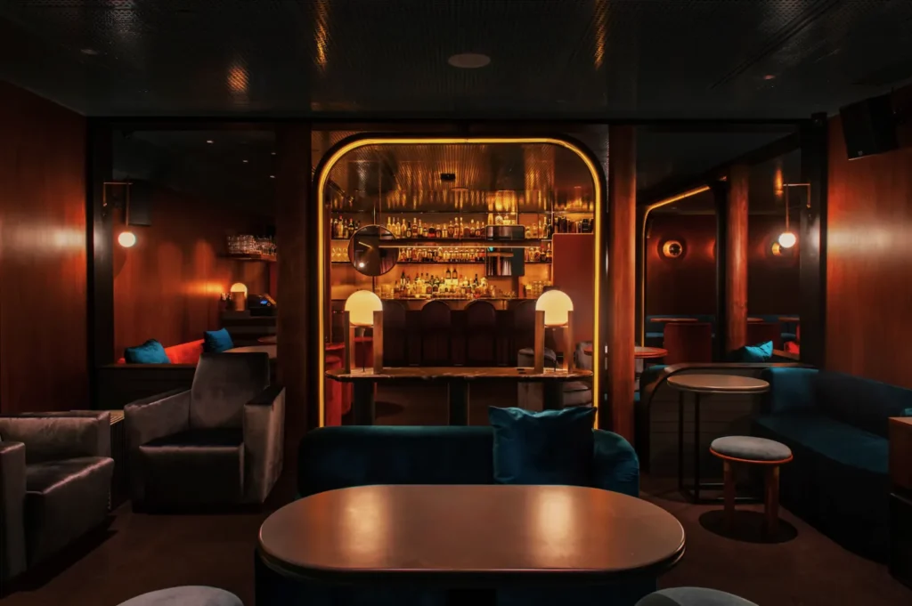 Chic lounge bar with warm lighting, plush blue seating, and wood-paneled walls. A backlit bar with various bottles adds an elegant, cozy ambiance.