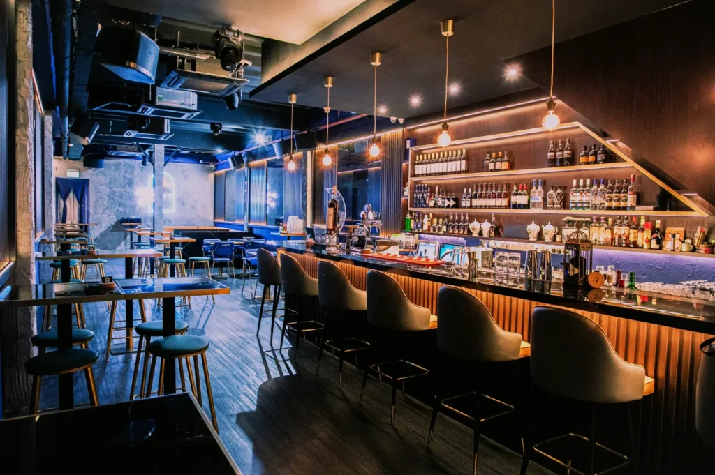 Stylish, dimly lit bar interior with a long counter, modern barstools, pendant lights, and shelves stocked with bottles. Cozy, elegant ambiance.